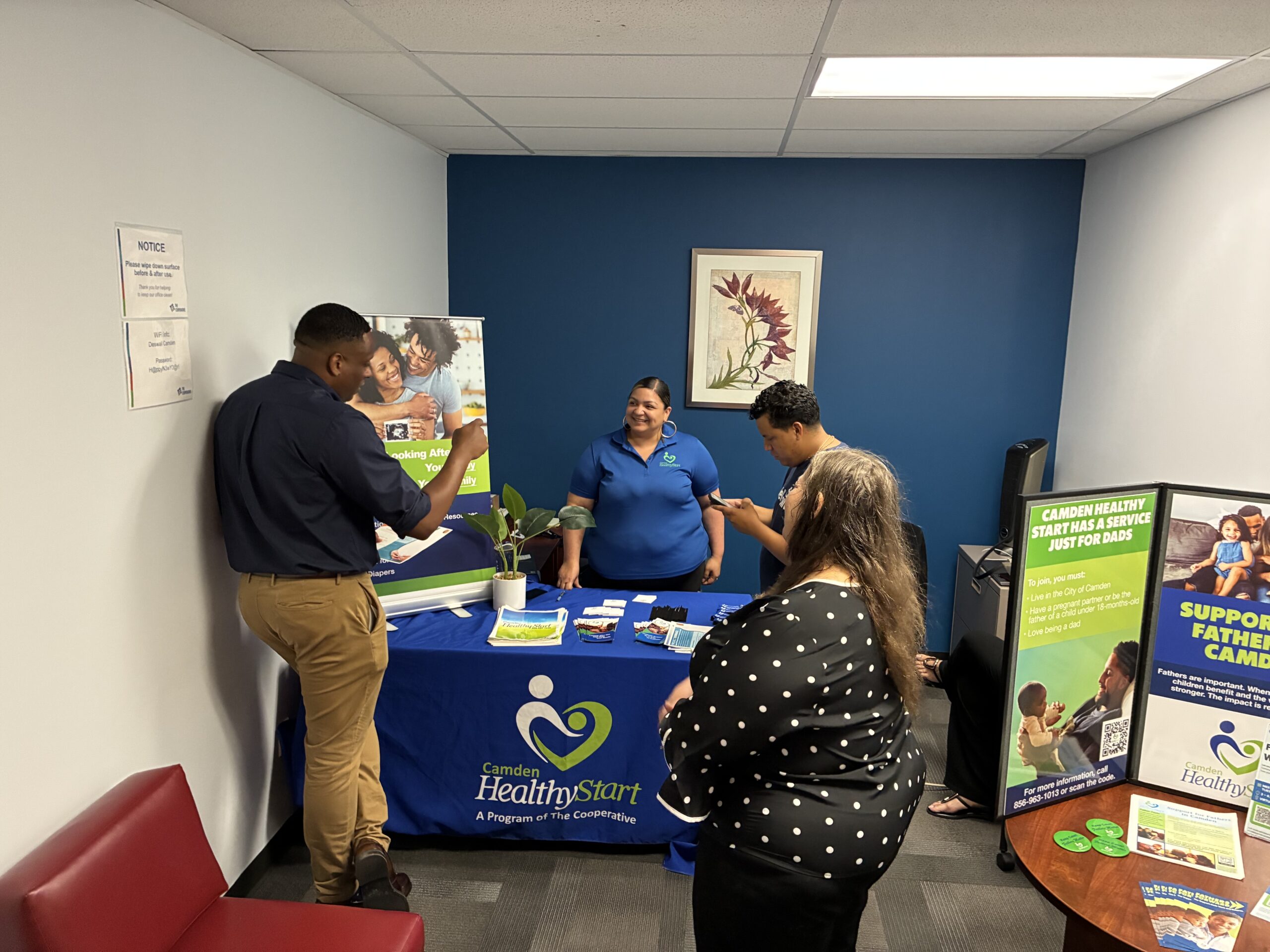 Smiling staff member standing behind a Camden Healthy Start information table while people look over resources.