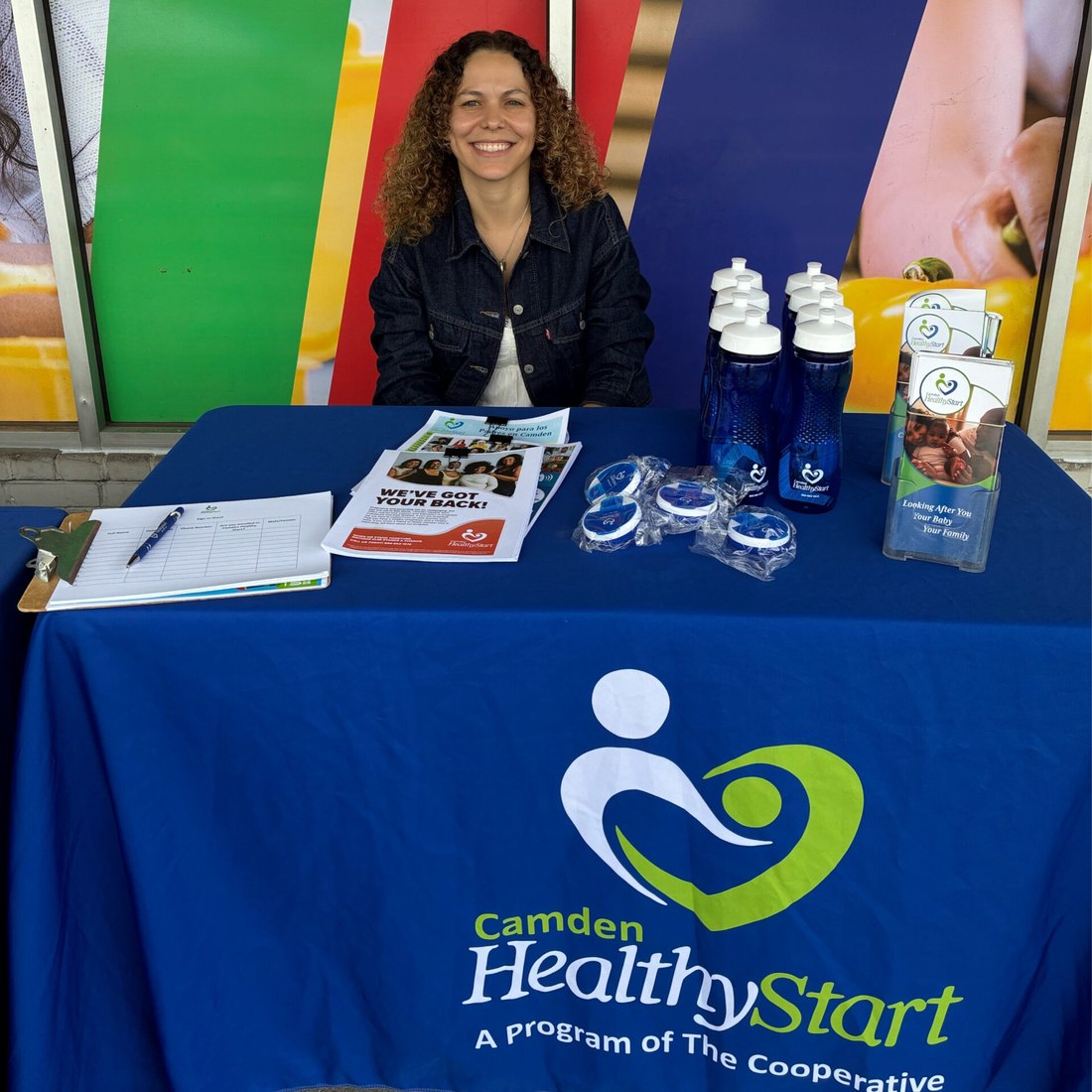A Camden Healthy Start staff member at a community resource table with program giveaways and the Camden Healthy Start tablecloth